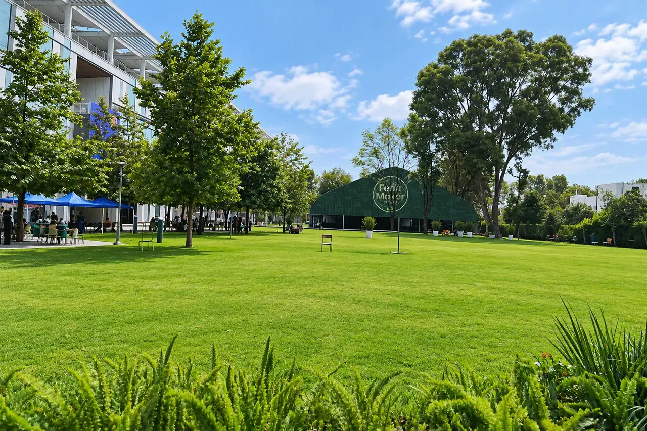 Jardín del campus con árboles, un edificio a la izquierda y, al fondo, una carpa verde con el logo de la Feria Maker